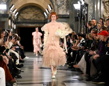 Model in layered pastel pink gown with floral elements walks runway, surrounded by audience, at Simone Rocha Spring/Summer 2023.