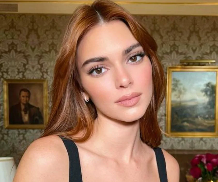 A woman with long brown hair poses indoors, with framed art on a wallpapered wall behind her.