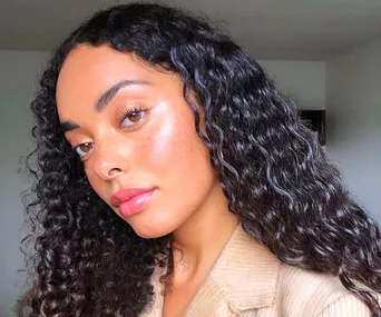 A woman with curly hair and glowing skin poses for a selfie, wearing a beige top, showcasing a natural makeup look.