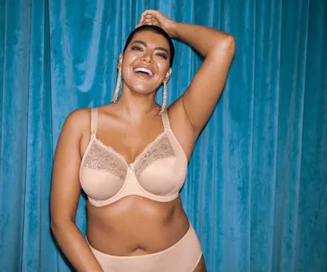 Model smiling in beige lingerie against a blue curtain background.