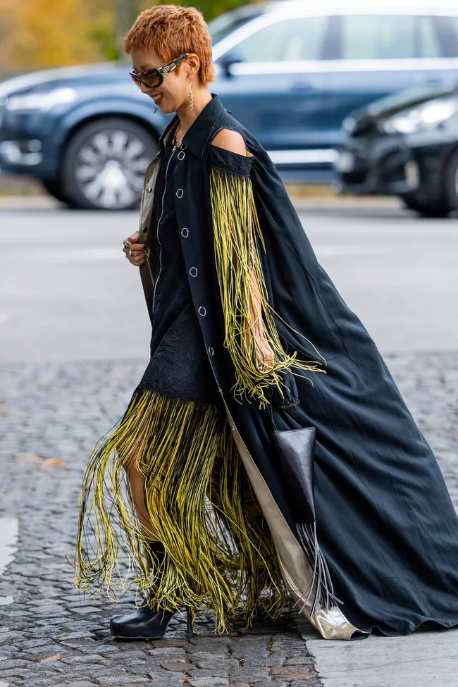 Woman in a black and yellow fringe dress with short hair and sunglasses, walking on the street.