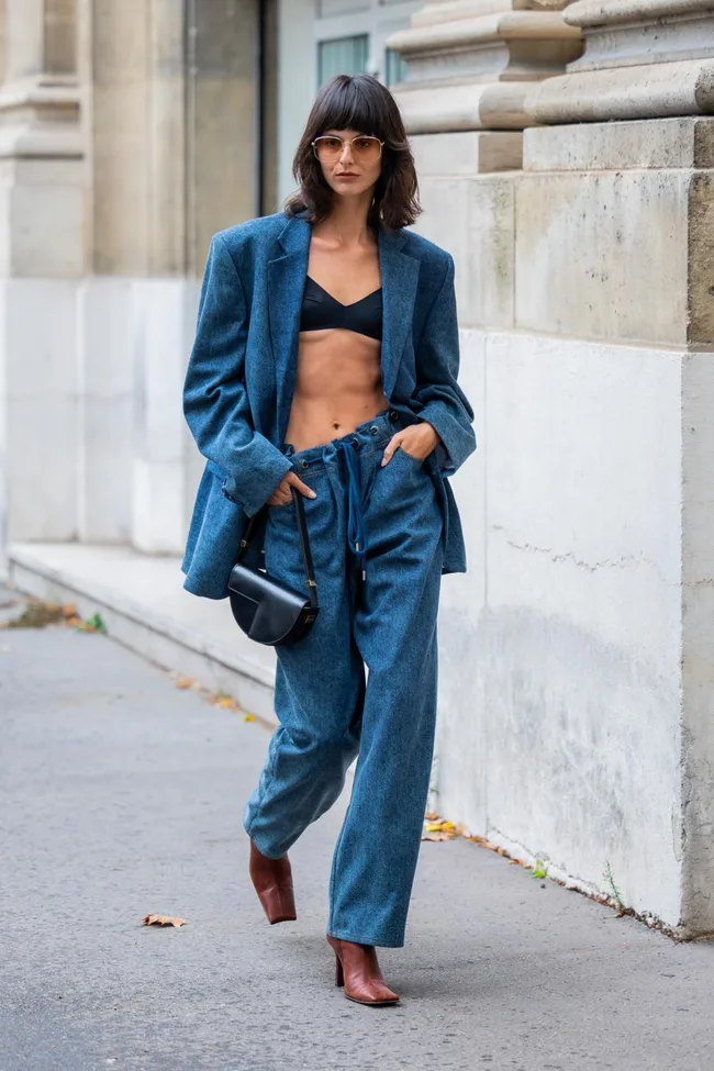 A woman walking in a blue suit, black bralette, and brown boots, carrying a black purse on a city street.