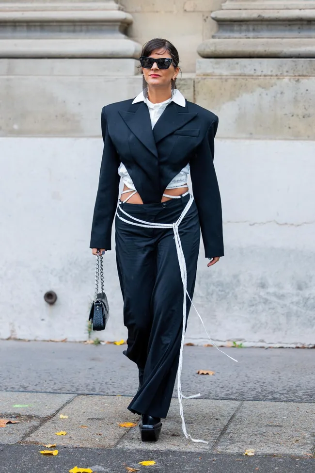 Person in a bold black suit with long white ties, sunglasses, and a small purse, walking on a city street.