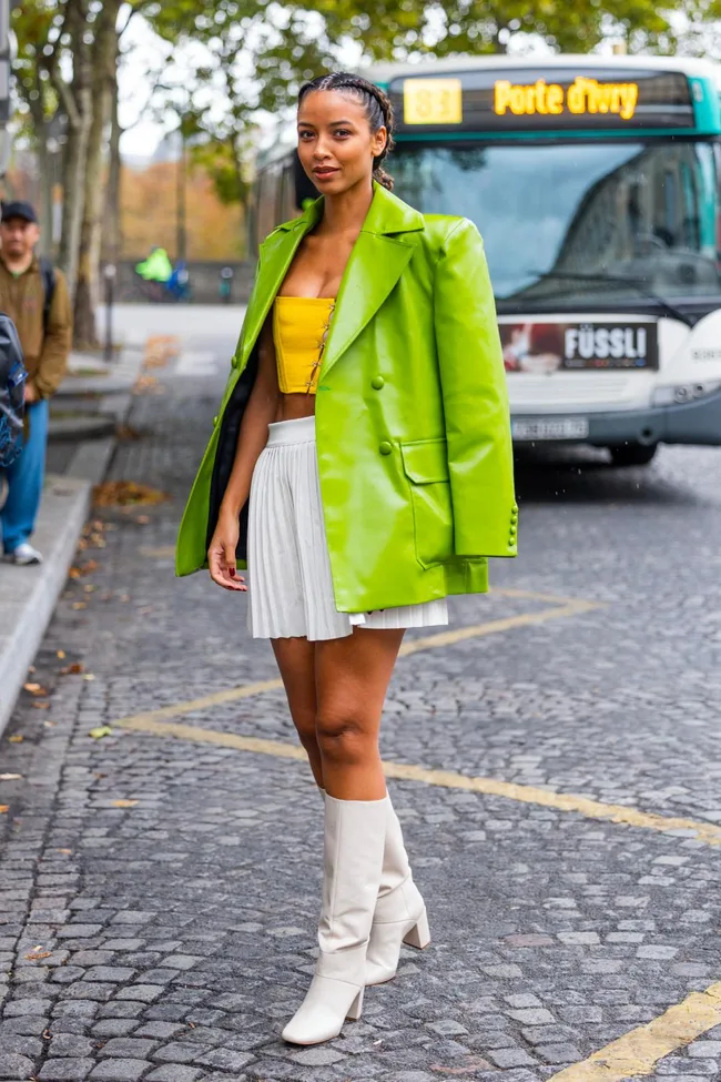 Street style: Woman in a green blazer, yellow top, white skirt, and boots, with braided hair, near a bus in Paris.