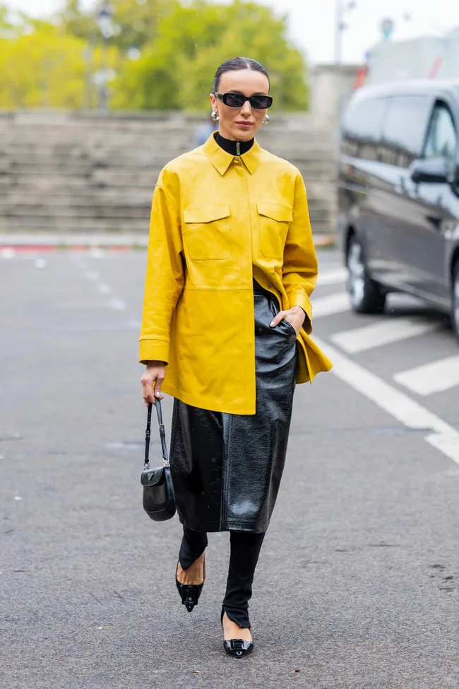 A person in a yellow jacket, black leather skirt, and sunglasses walking on a street with a car and trees in the background.