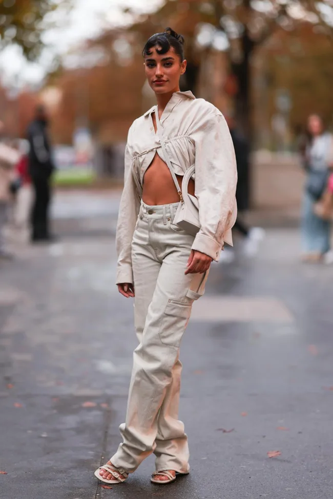 A woman in a stylish beige outfit poses confidently on a city street, with autumn trees in the background.