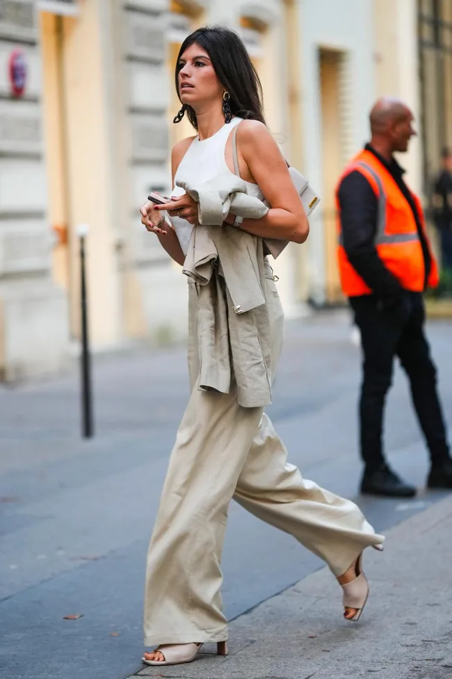 Woman in a sleeveless top and wide-leg pants walking on the street, holding a phone and jacket.