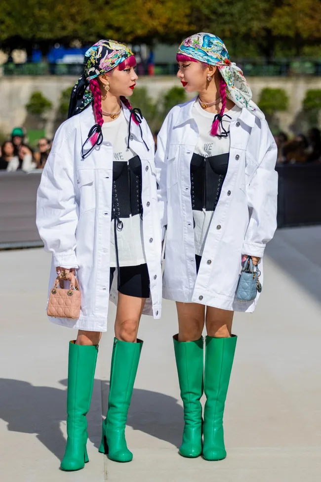 Two women with pink braids and headscarves in matching white jackets and green boots, holding handbags.