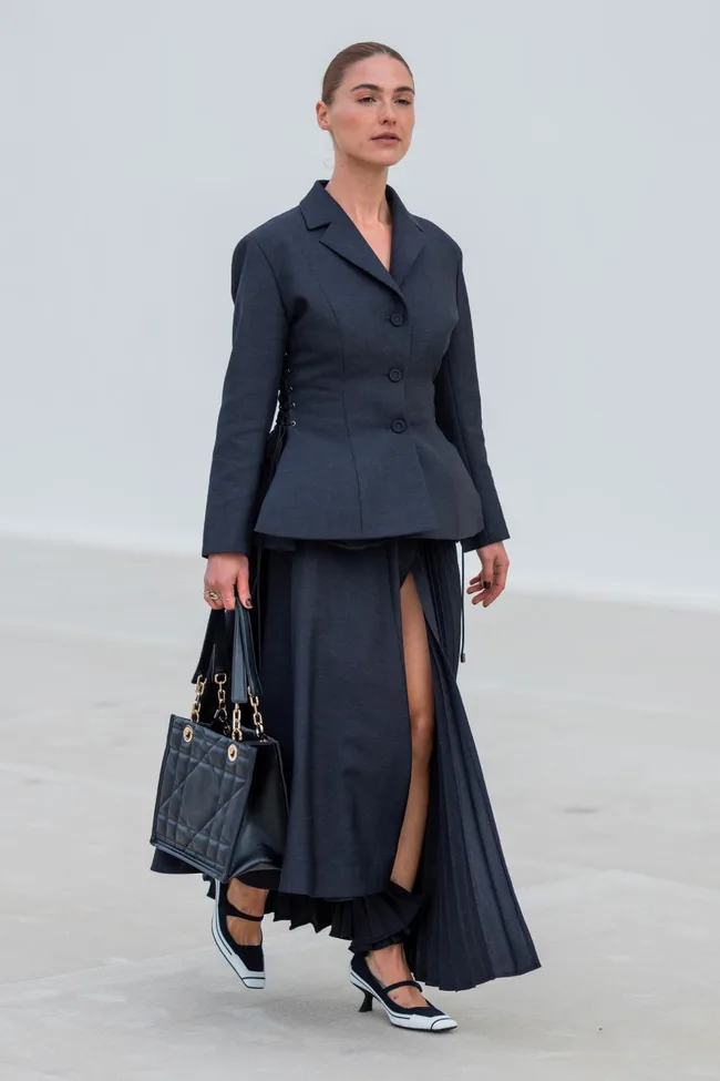 A model wearing a dark tailored blazer and skirt, carrying a black handbag and walking on a light surface.