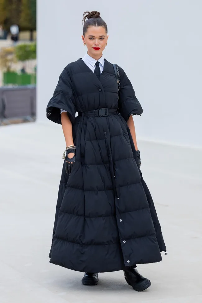 Woman in long black padded dress with a belted waist, white shirt, and tie, standing outdoors.