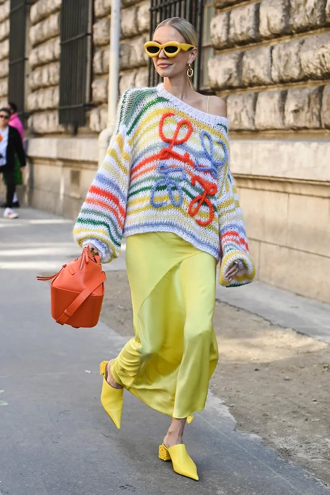 Stylish woman in a colorful sweater, yellow skirt, and sunglasses, holding an orange bag, walking on street.