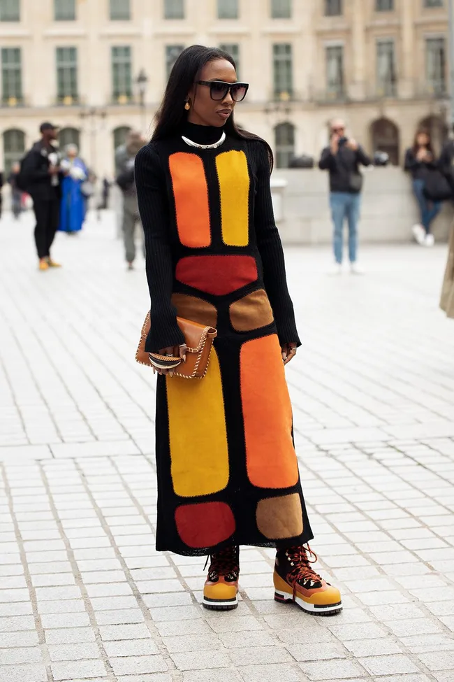 Alt text: Woman in colorful geometric dress with sunglasses and holding a brown bag, standing on a city street.
