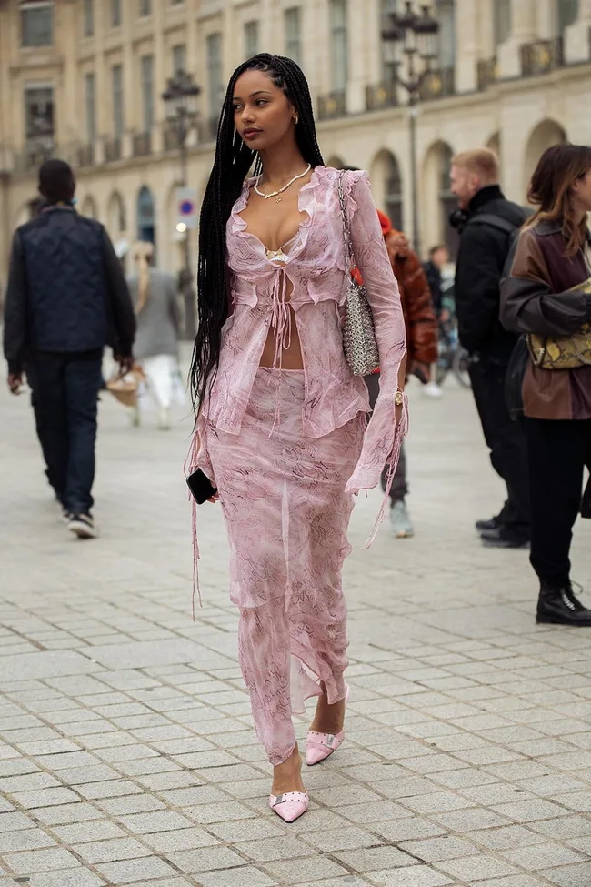 Young woman in pink lace outfit with long braids walks on cobblestone street, carrying a handbag and phone.