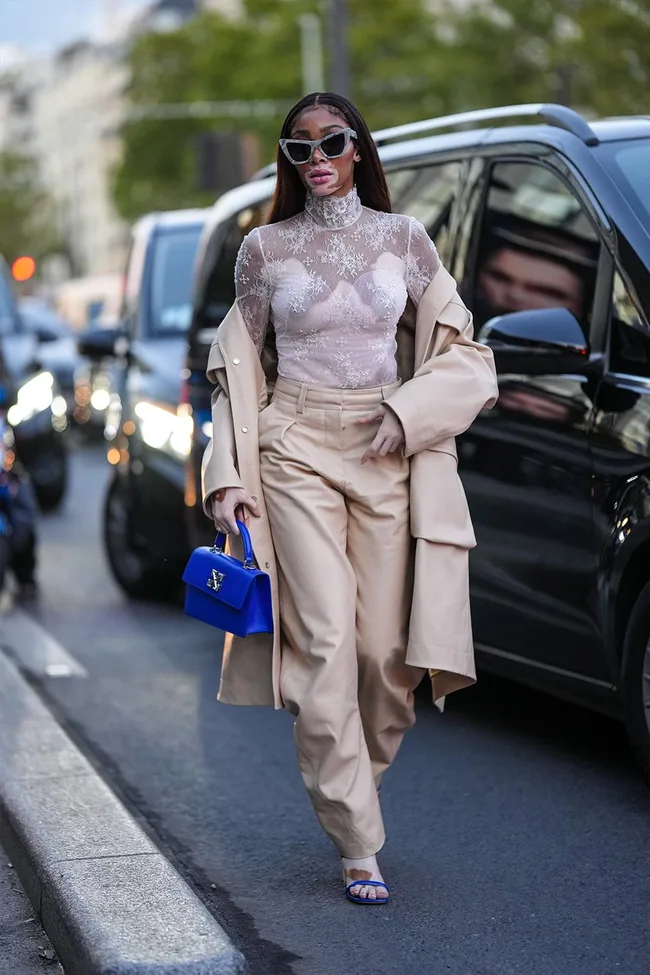 Woman in sheer lace top, beige trench coat, wide-leg pants, blue handbag, and sunglasses walking on a street.