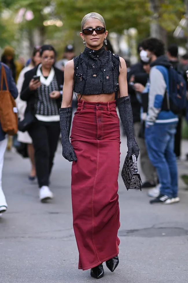 A woman in a black crop top and red skirt walks on a street, wearing sunglasses and gloves, carrying a textured handbag.