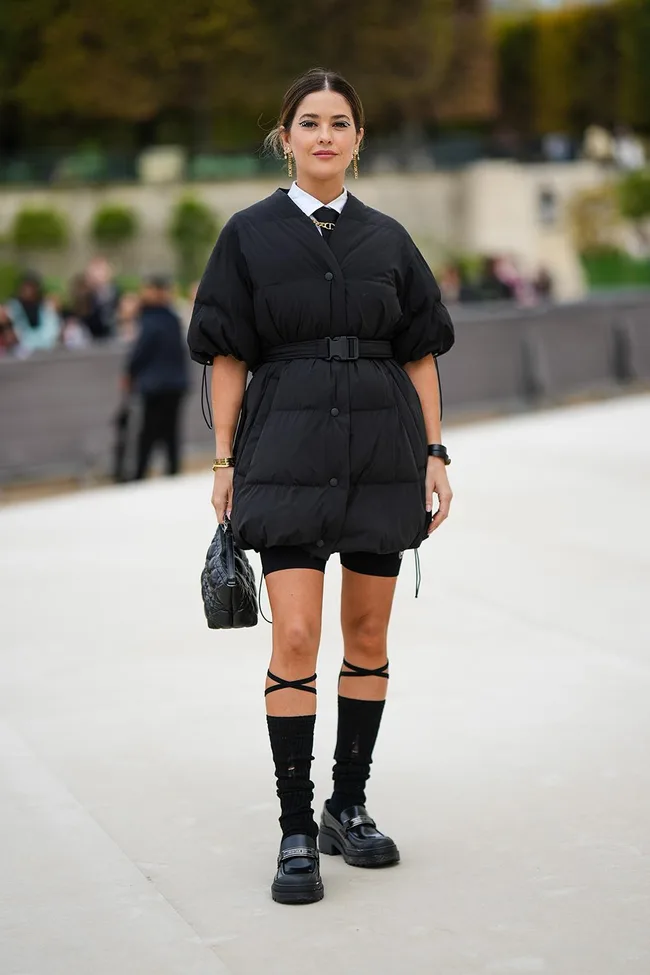 A woman in a black belted jacket dress with knee-high socks and chunky shoes stands outdoors.
