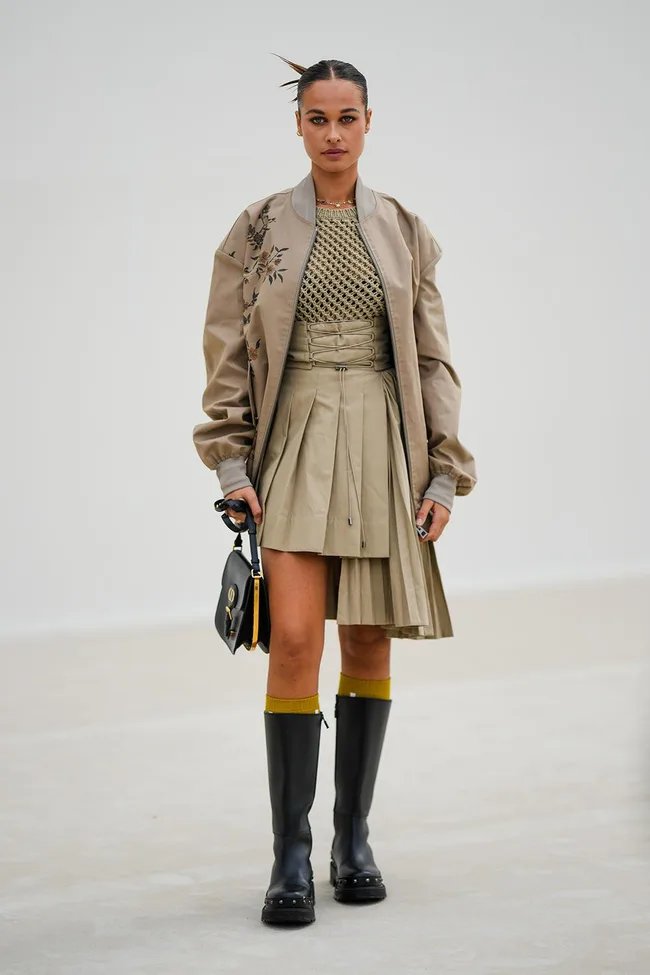 Woman in a beige jacket and skirt, holding a black handbag, wearing knee-high black boots on a plain background.