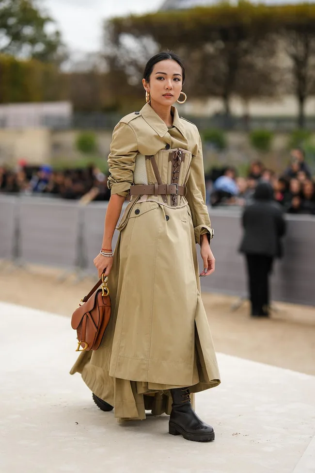 Woman in a tan trench coat with a brown handbag and hoop earrings, walking outdoors.