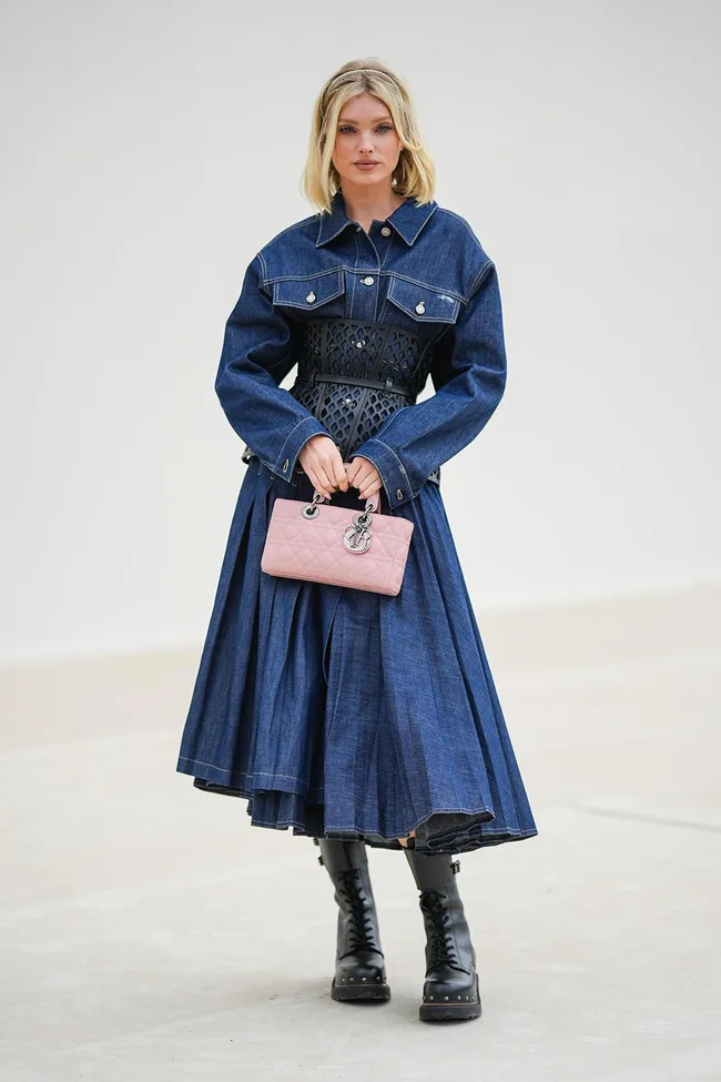 Woman in denim dress with a black corset, holding a pink purse, wearing black boots.