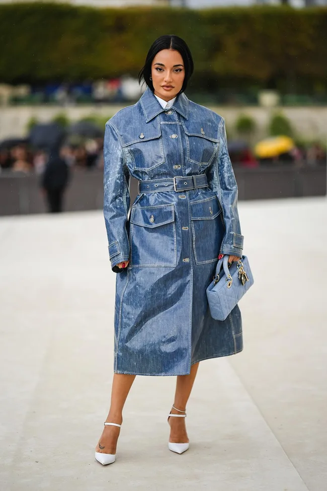 Person in a blue denim trench coat and white heels, holding a matching blue purse, standing outside.