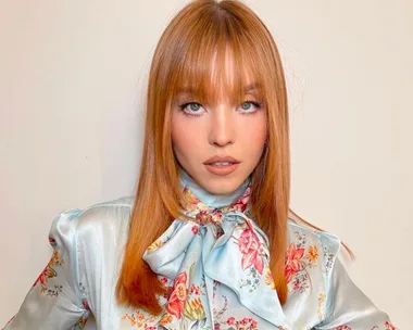 Woman with straight reddish hair and bangs, wearing a floral silk blouse, poses against a neutral background.