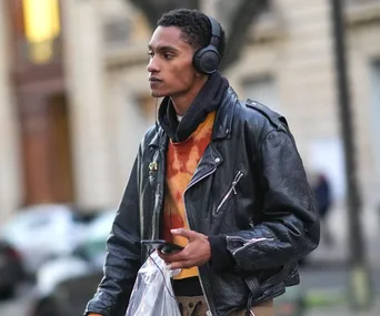 A person with headphones, wearing a leather jacket and holding a plastic bag, walking outdoors.