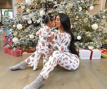 A mother and child in matching Christmas pajamas sit and kiss by a decorated tree with gifts underneath.