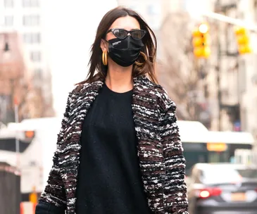 Woman in urban setting wearing a black face mask, sunglasses, large hoop earrings, dark sweater, and patterned coat.