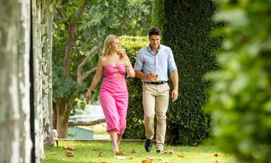 A couple walks on grass, holding drinks, surrounded by lush green trees and a hedge.