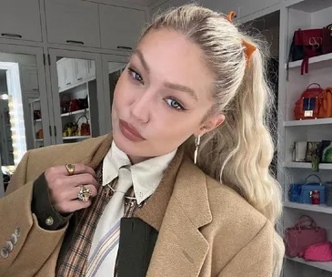 A woman with blond hair in a ponytail, wearing a beige coat, plaid shirt, and tie, stands in a handbag-filled closet.