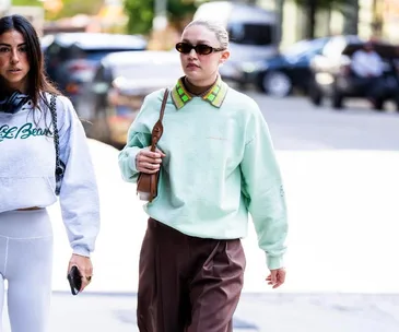 A person wearing a mint green sweater and brown pants walks on a street, accompanied by another person in casual attire.