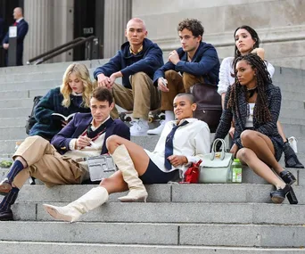 Seven young adults in stylish outfits sit on steps, engaged in conversation and relaxing, from "Gossip Girl" reboot.