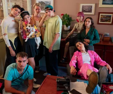 A group of young, stylishly dressed people pose casually in an office setting, with vibrant clothing and a dog.