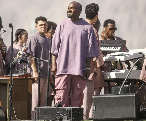 Man in pink-purple outfit performs on stage with musicians, surrounded by instruments and a beige backdrop.