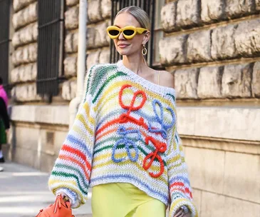 Woman in colorful striped sweater with floral details, yellow sunglasses and skirt, holding an orange handbag, street style.