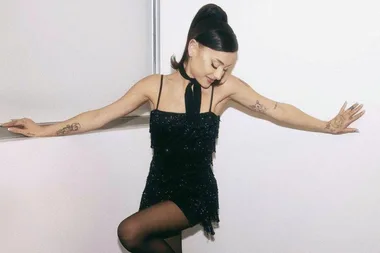 Ariana Grande in a sparkling black mini dress, black stockings, with tattoos visible and hair styled in a ponytail.