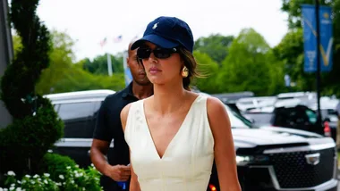 Woman in a white dress wearing a navy cap and sunglasses, walking outdoors with cars and greenery in the background.