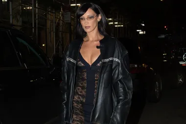 A woman in a black lace dress and leather jacket walks at night, with scaffolding and cars in the background.