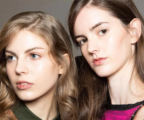 Two young women with clear skin and natural makeup, looking to the side against a neutral background.