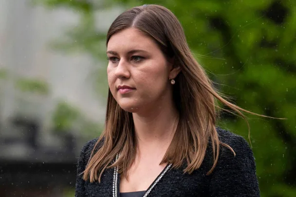 A person with long brown hair stands outdoors with a neutral expression, dressed in a dark jacket.