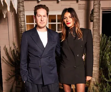 A man and woman in tailored suits stand together, posing in a warmly lit outdoor setting.