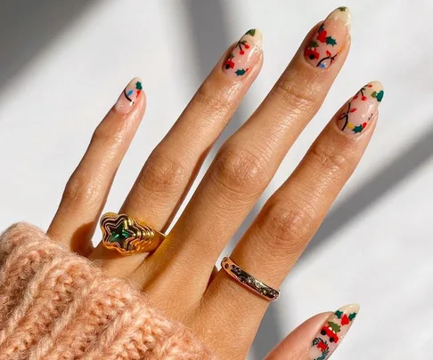 Hand with festive nail art and two rings, one gold with a green stone, wearing a pink sweater.