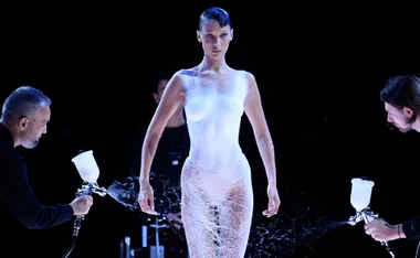 Model standing as two people spray a white dress onto her body during a fashion show.