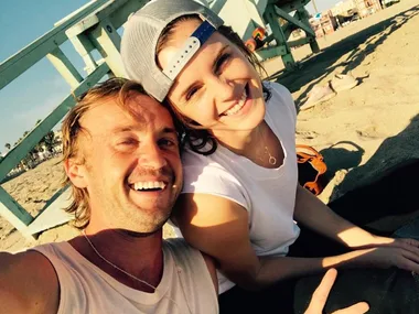 Two people smiling at the camera on a sunny beach, with a lifeguard stand in the background.