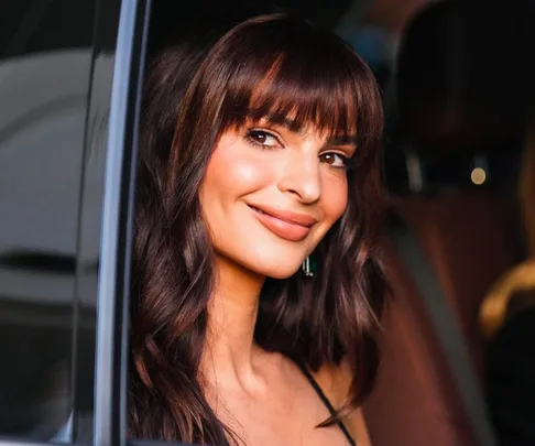 A woman with long brown hair and bangs smiles from a car window.