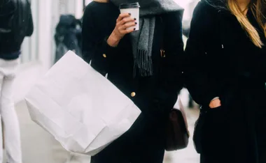 Two people walking, one holding a large white shopping bag and a coffee cup, dressed in dark coats and scarves.