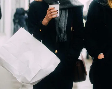 Two people walking, one holding a large white shopping bag and a coffee cup, dressed in dark coats and scarves.