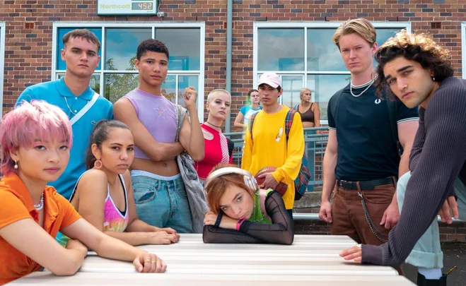 Group of teens at a school, one with pink hair and another leaning on table with headphones. Scene from "Heartbreak High".