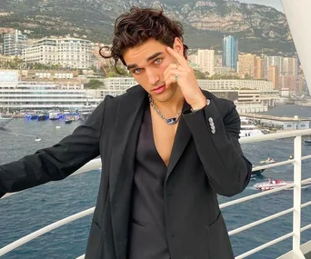 Man in black suit poses on a balcony overlooking a harbor with cityscape and mountains in the background.