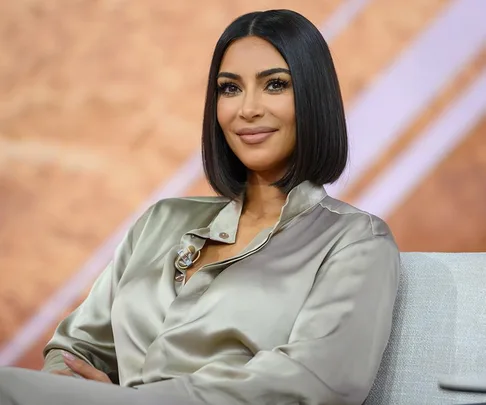 Kim Kardashian sitting on a couch, wearing a silk blouse, with a neutral expression.